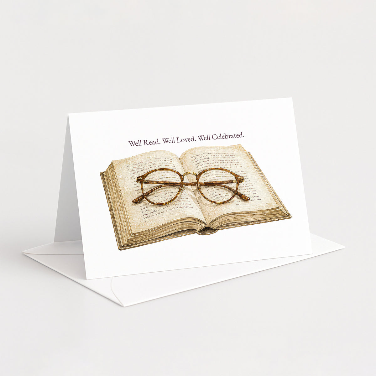 A white greeting card standing upright on a white envelope, printed with an illustration of an open book and round eyeglasses, with the text 'Well Read. Well Loved.