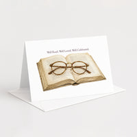 A white greeting card standing upright on a white envelope, printed with an illustration of an open book and round eyeglasses, with the text 'Well Read. Well Loved.