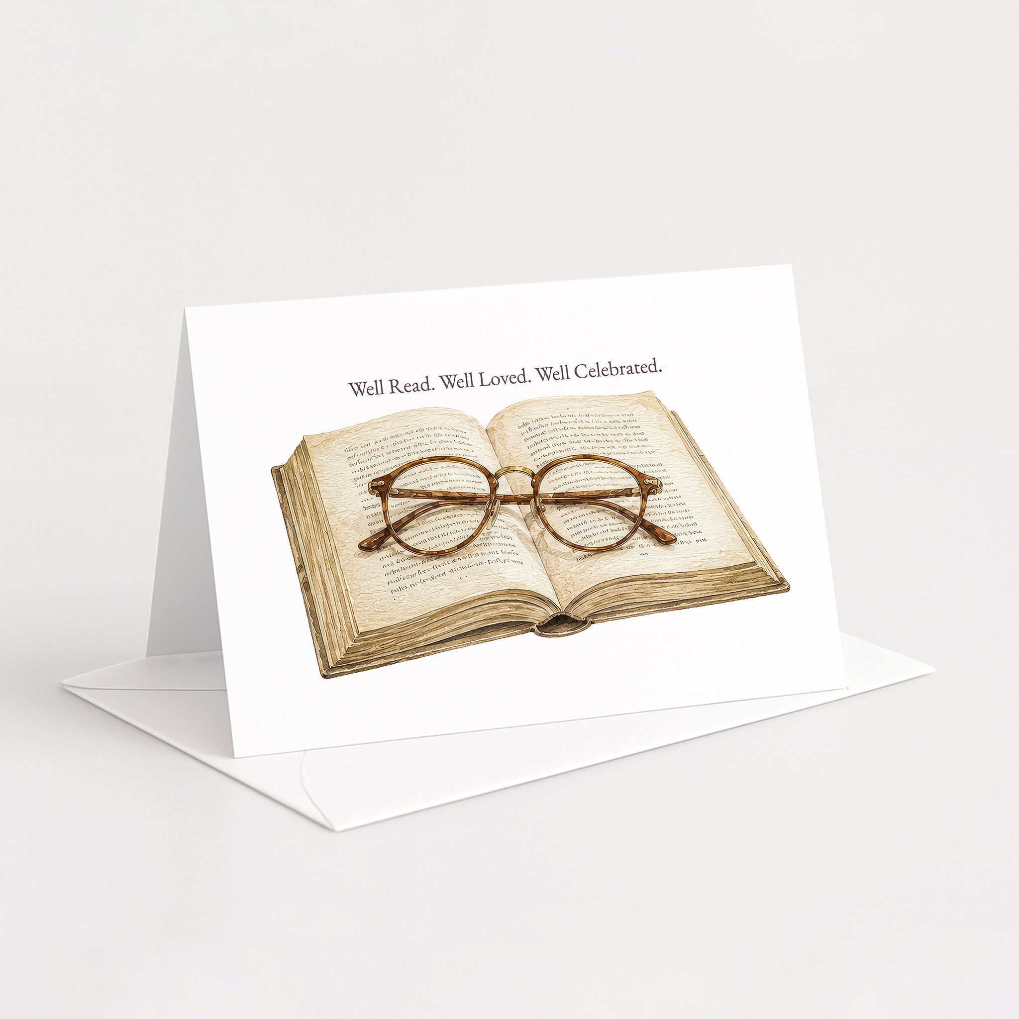 A white greeting card standing upright on a white envelope, printed with an illustration of an open book and round eyeglasses, with the text 'Well Read. Well Loved.