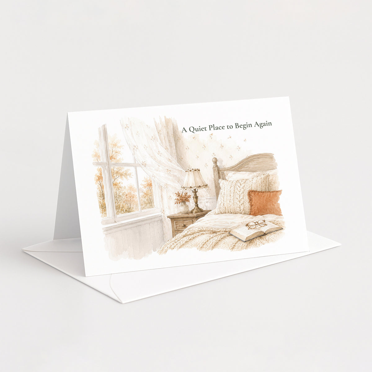 A folded greeting card standing open on a white envelope against a light gray background. The front of the card shows a soft watercolor illustration of a bedroom scene with a bed, window, curtains, and the text 'A Quiet Place to Begin Again'.