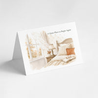 A folded greeting card standing on its front flap against a light gray background. The card features a watercolor-style illustration of a cozy bedroom scene with a bed, window, curtains, and the text 'A Quiet Place to Begin Again' printed near the top.