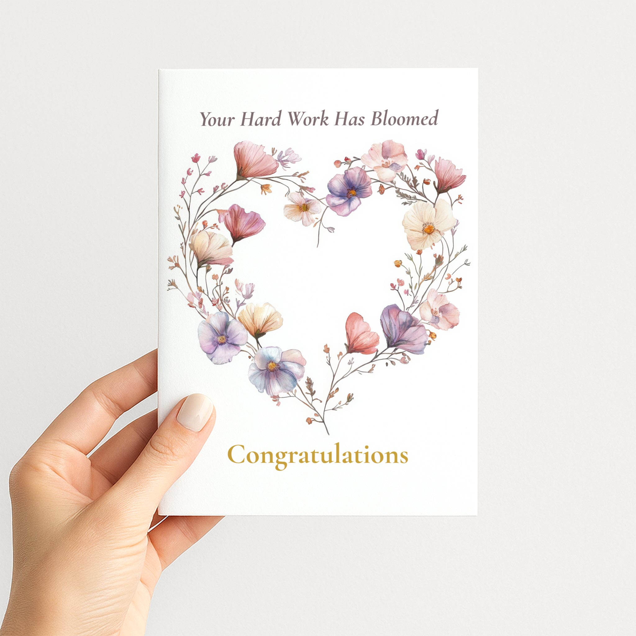 Hand holding a white congratulations card decorated with a heart-shaped watercolor wildflower wreath.