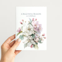 Hand holding A Beautiful Reason to Celebrate card featuring pink and white botanical artwork.