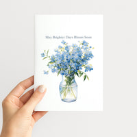 Hand holding a white greeting card illustrated with blue flowers in a glass vase and the words “May Brighter Days Bloom Soon.”