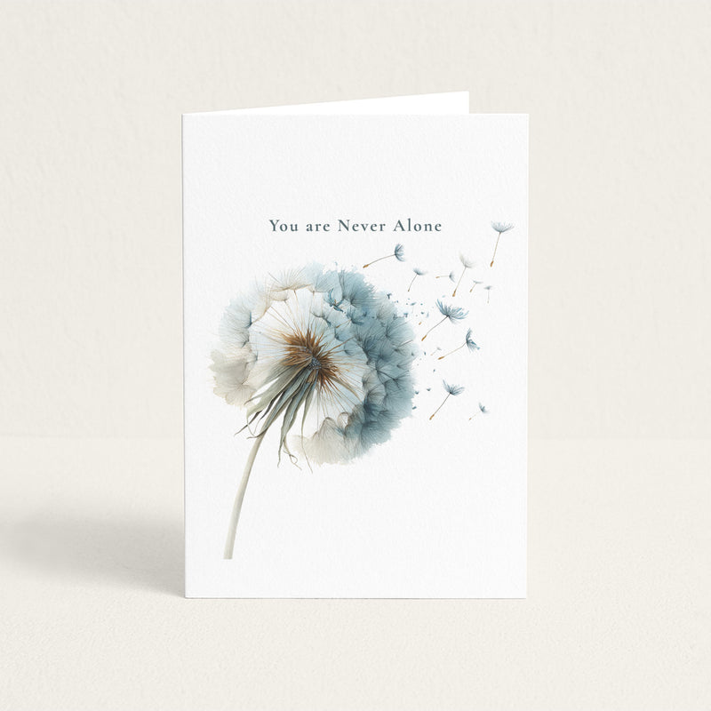 Encouragement card featuring a soft blue watercolor dandelion with seeds drifting away beneath the words “You are Never Alone.”