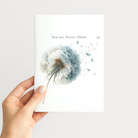 Hand holding a white greeting card illustrated with a pale blue dandelion and floating seeds.