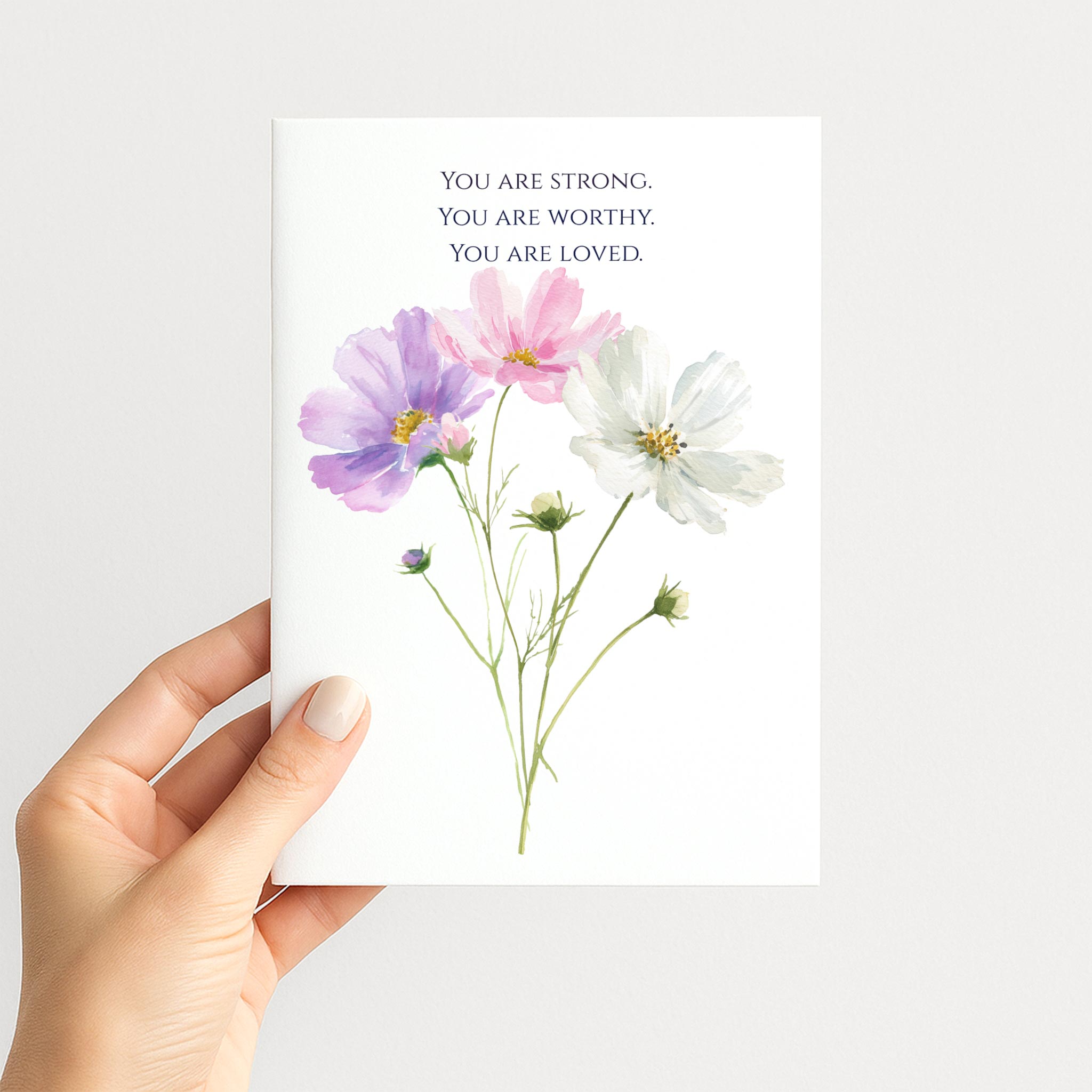 Hand holding a white encouragement card illustrated with delicate pink, lavender, and white watercolor flowers.