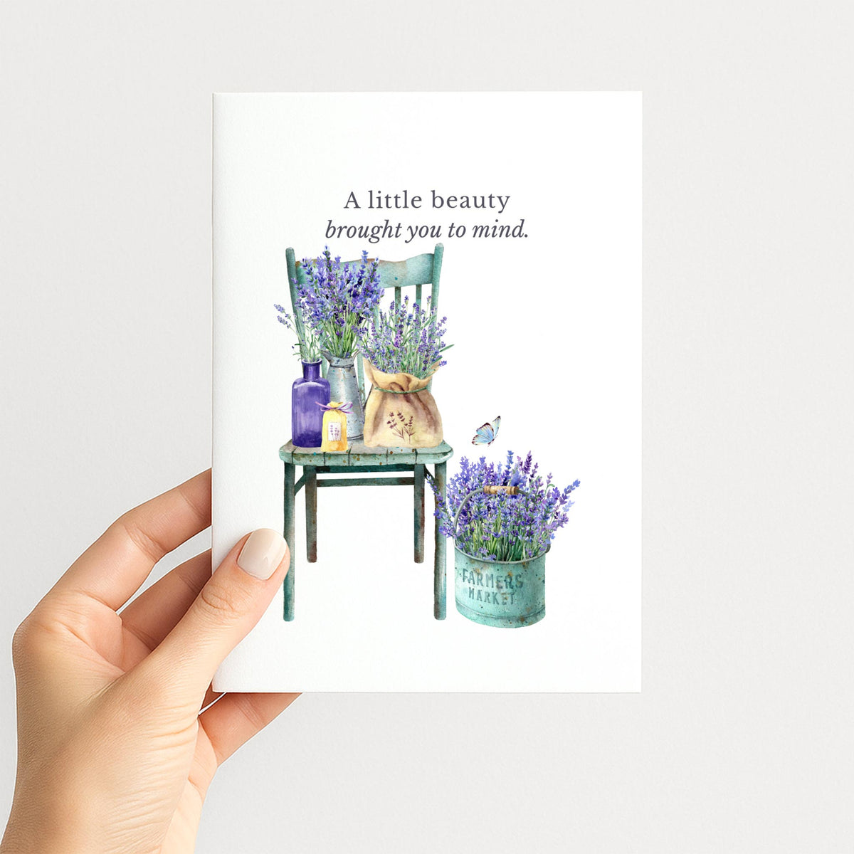 Hand holding a greeting card illustrated with lavender bouquets, a vintage green chair, a market pail, and a small butterfly.
