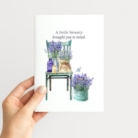 Hand holding a greeting card illustrated with lavender bouquets, a vintage green chair, a market pail, and a small butterfly.