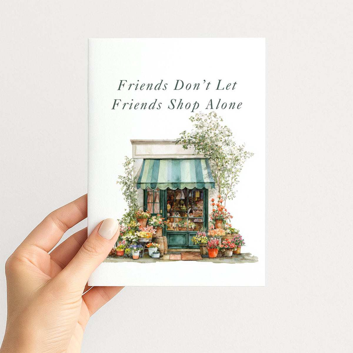 Hand holding a friendship card featuring a charming watercolor flower shop surrounded by colorful blooms.