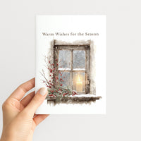 Hand holding a winter greeting card illustrated with a candle glowing in a snowy window framed by evergreen branches and red berries.