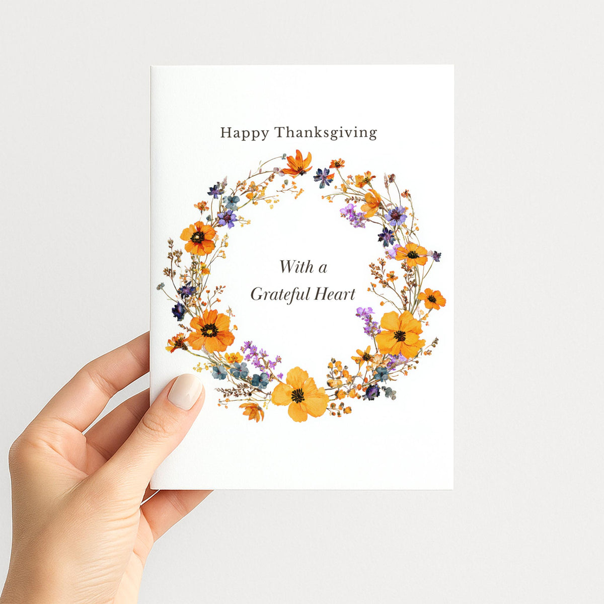Hand holding a Thanksgiving card with an orange and purple autumn floral wreath and “With a Grateful Heart.”