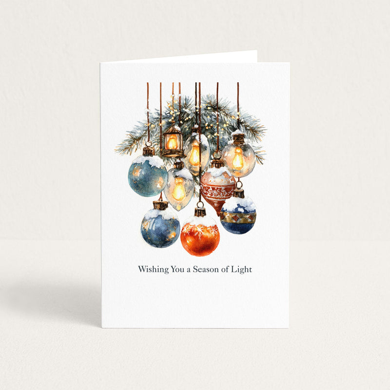 Wishing You a Season of Light holiday card featuring watercolor ornaments, glowing lanterns, and snowy evergreen branches.