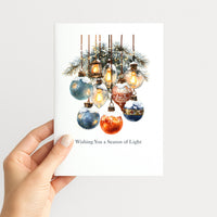 Hand holding a holiday card featuring blue and red ornaments, glowing lights, and snow-covered greenery.