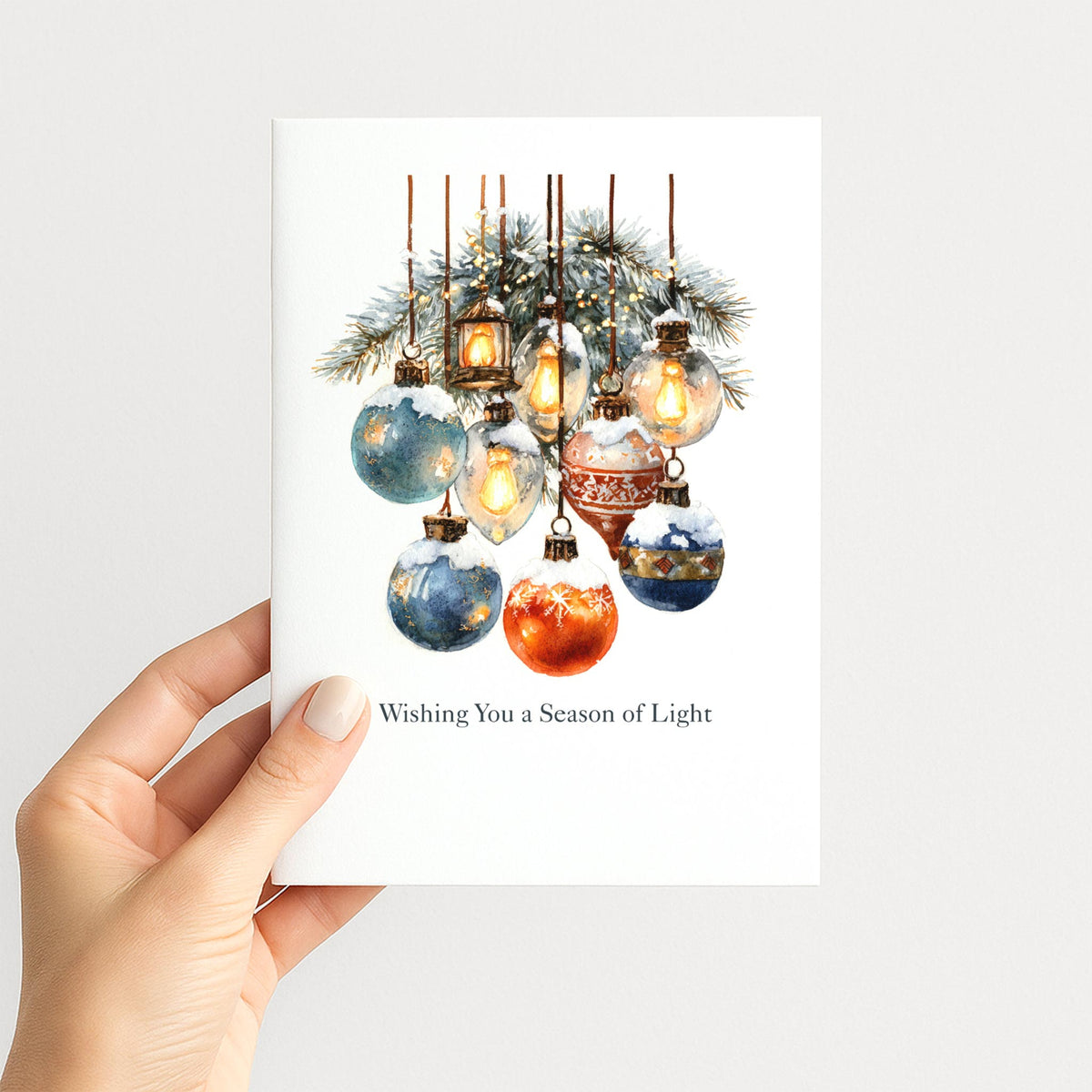 Hand holding a holiday card featuring blue and red ornaments, glowing lights, and snow-covered greenery.