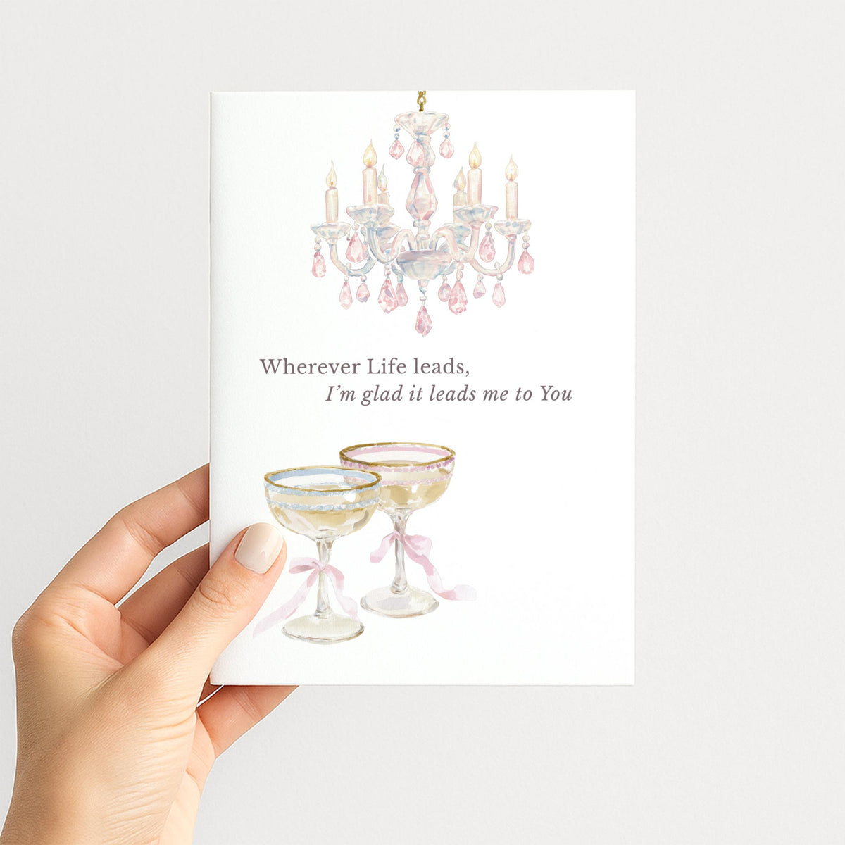 Hand holding a romantic greeting card illustrated with a pink crystal chandelier and two elegant champagne glasses tied with ribbons.