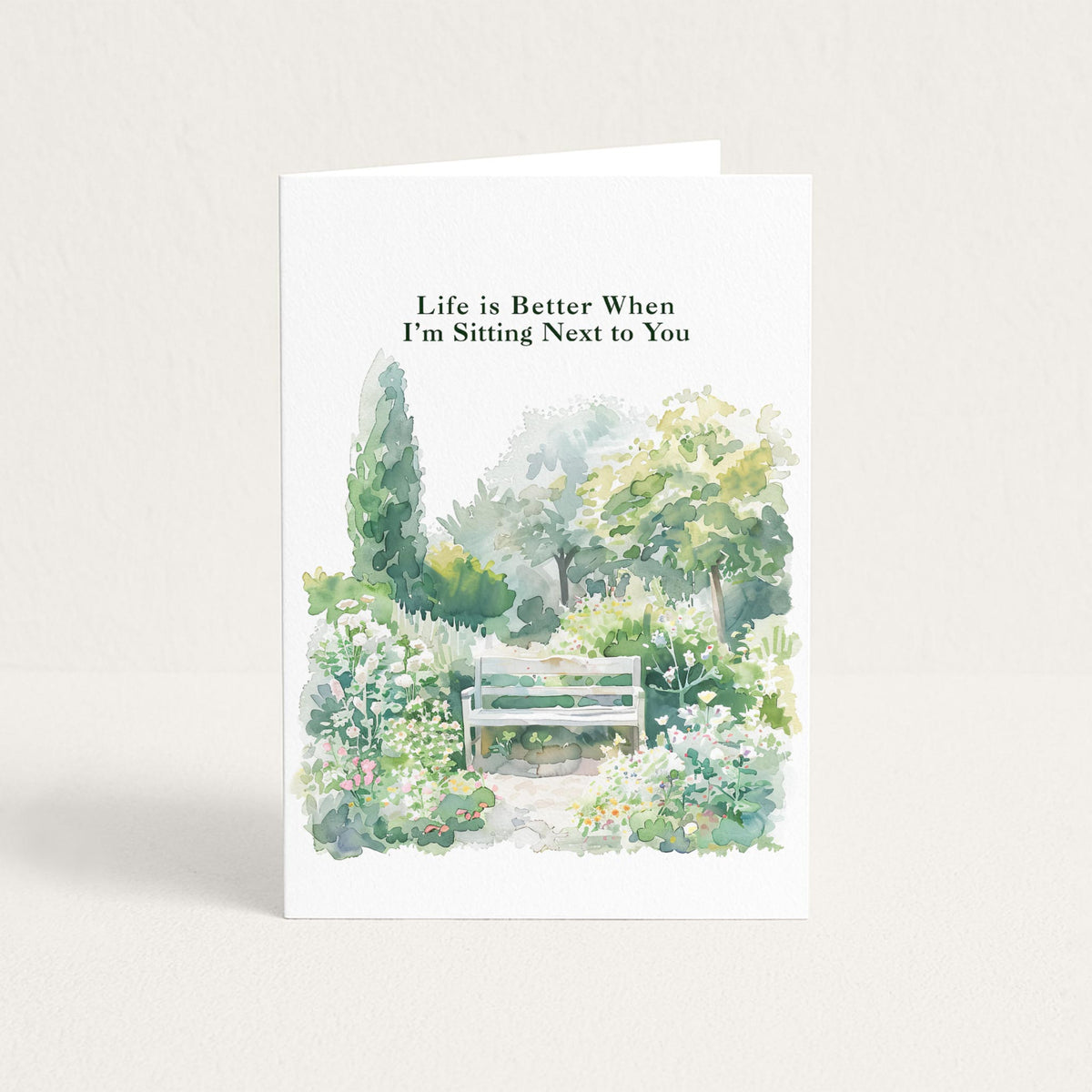 Love greeting card featuring a watercolor garden bench surrounded by soft green foliage and flowers with “Life is Better When I’m Sitting Next to You.”