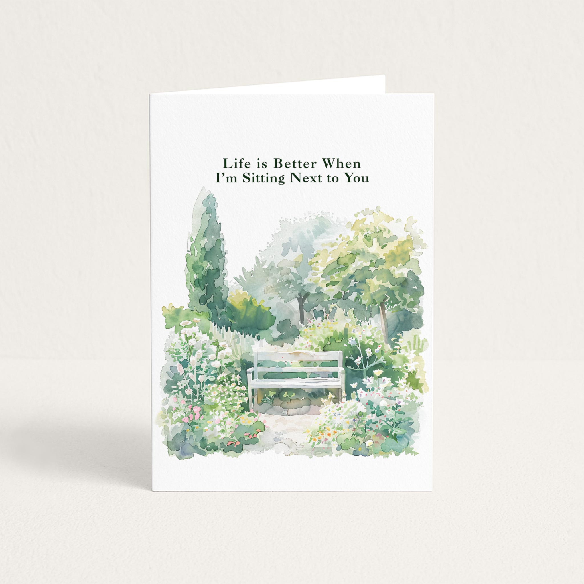 Love greeting card featuring a watercolor garden bench surrounded by soft green foliage and flowers with “Life is Better When I’m Sitting Next to You.”