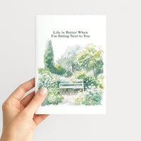 Hand holding a romantic greeting card illustrated with a peaceful flower garden and bench.
