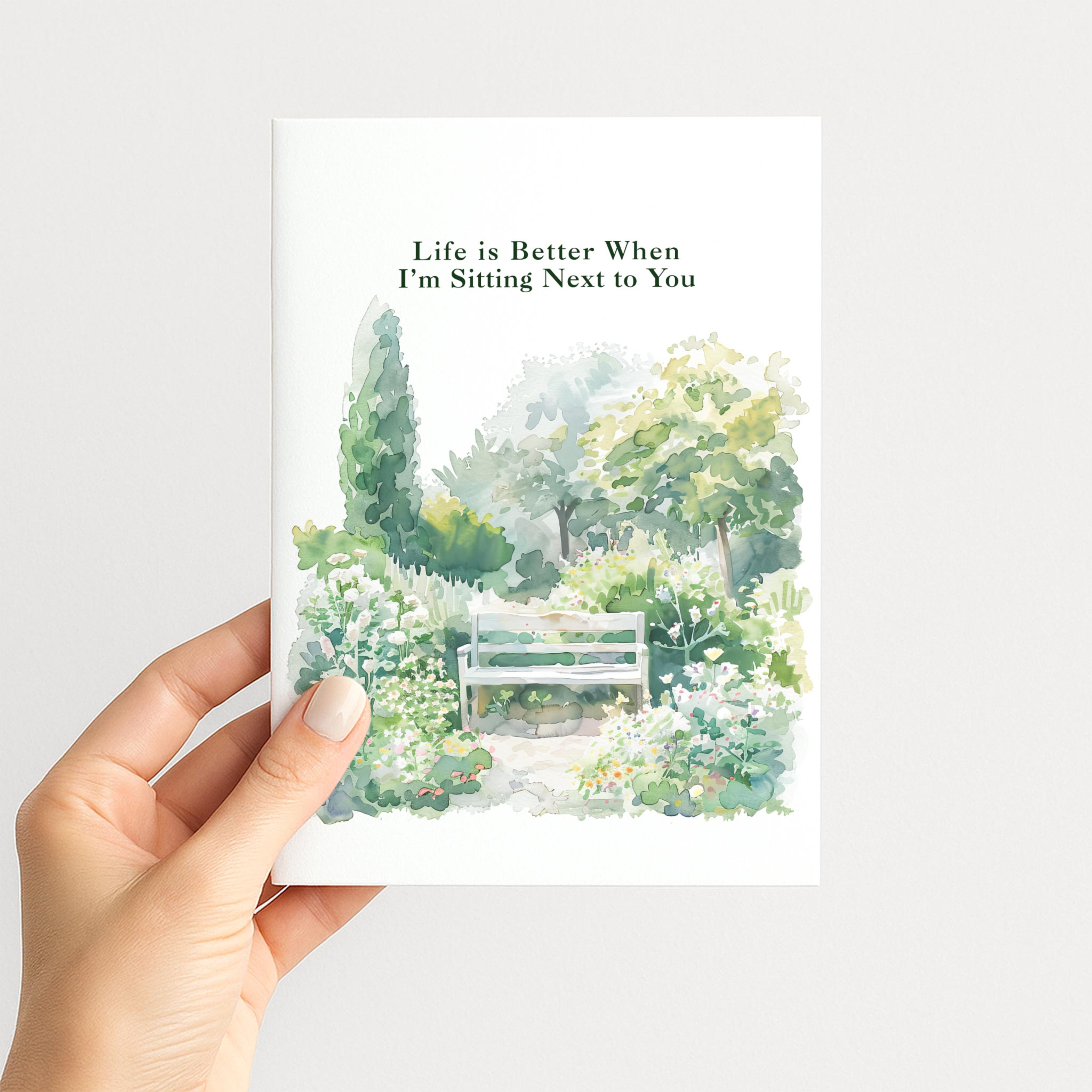 Hand holding a romantic greeting card illustrated with a peaceful flower garden and bench.