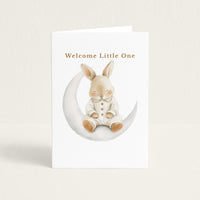 New baby greeting card featuring a watercolor bunny in striped pajamas seated on a crescent moon beneath the words “Welcome Little One.”
