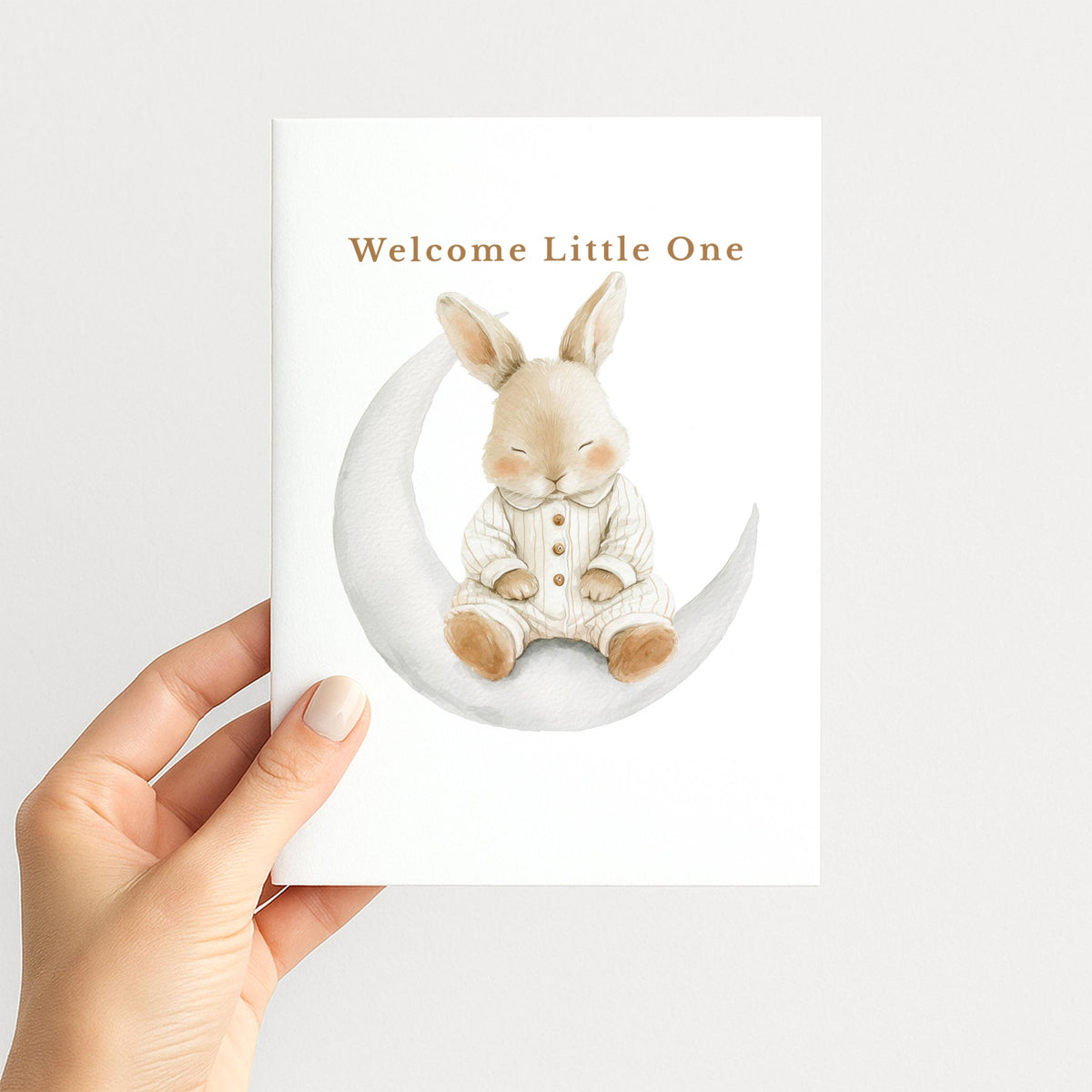 Hand holding a white new baby card illustrated with a sleepy watercolor bunny seated on a crescent moon.