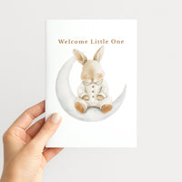 Hand holding a white new baby card illustrated with a sleepy watercolor bunny seated on a crescent moon.