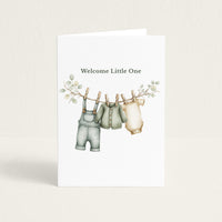 New baby greeting card featuring sage green overalls, a cardigan, and a cream bodysuit hanging from a leafy clothesline beneath the words “Welcome Little One.”