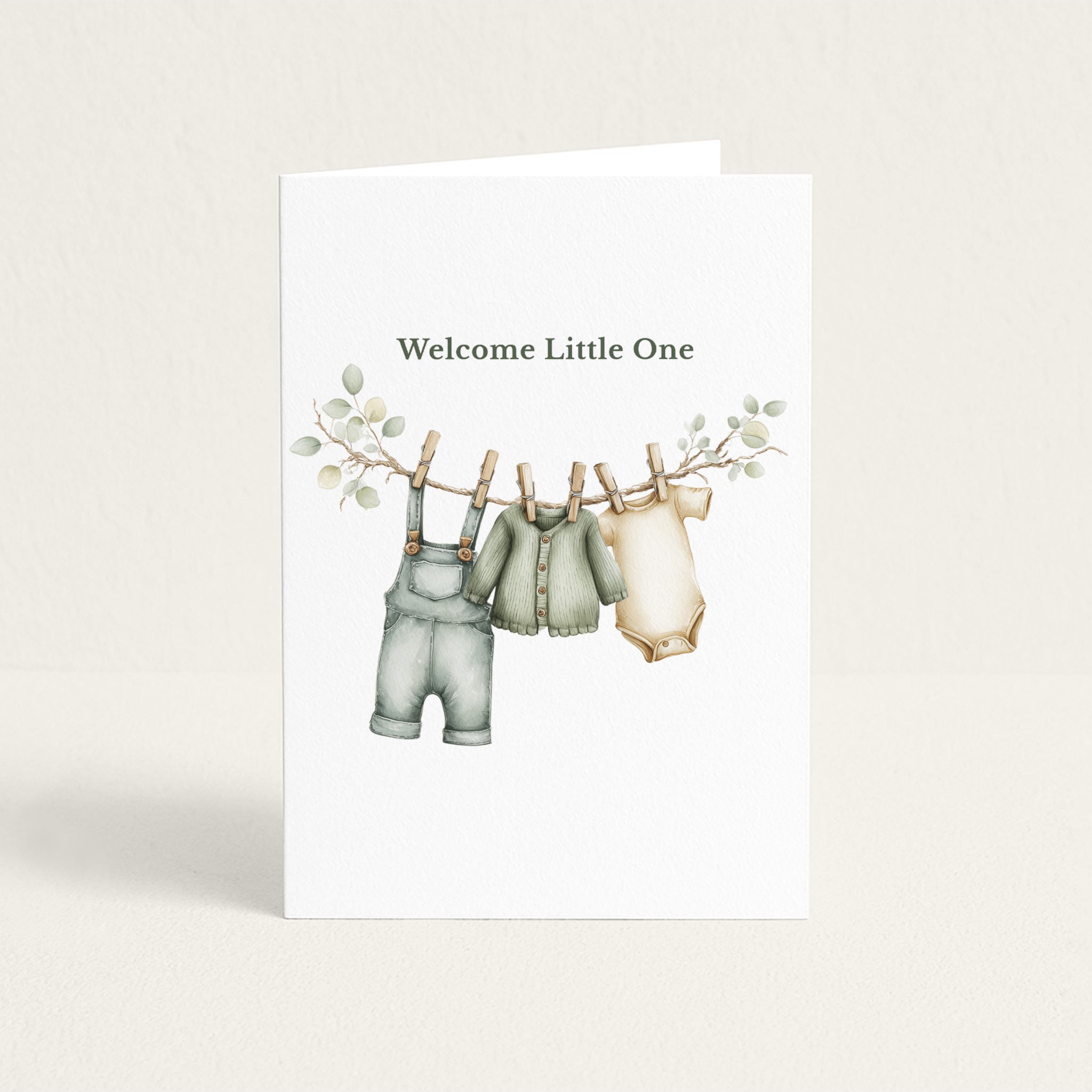 New baby greeting card featuring sage green overalls, a cardigan, and a cream bodysuit hanging from a leafy clothesline beneath the words “Welcome Little One.”