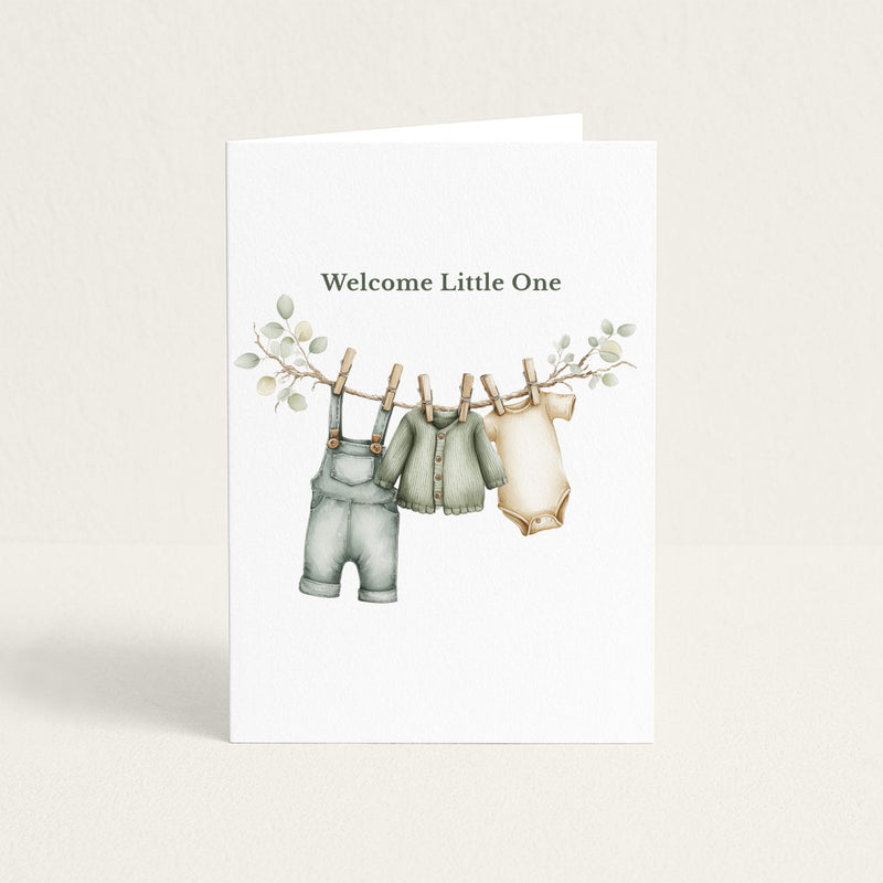 New baby greeting card featuring sage green overalls, a cardigan, and a cream bodysuit hanging from a leafy clothesline beneath the words “Welcome Little One.”