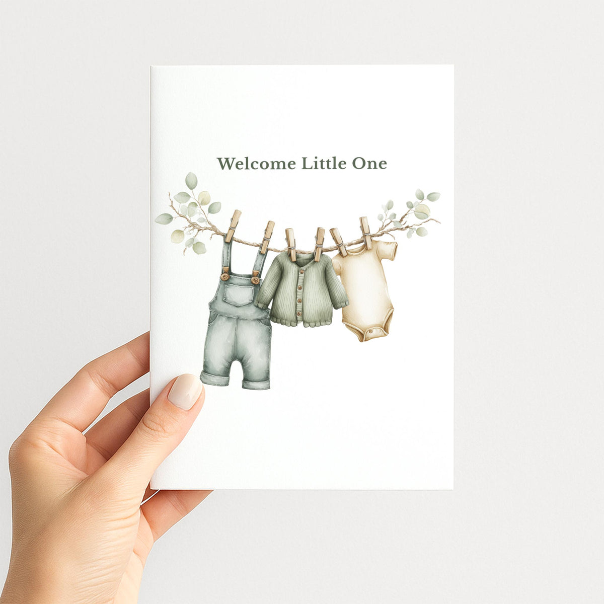 Hand holding a white new baby card illustrated with neutral baby clothes hanging from a eucalyptus-trimmed clothesline.