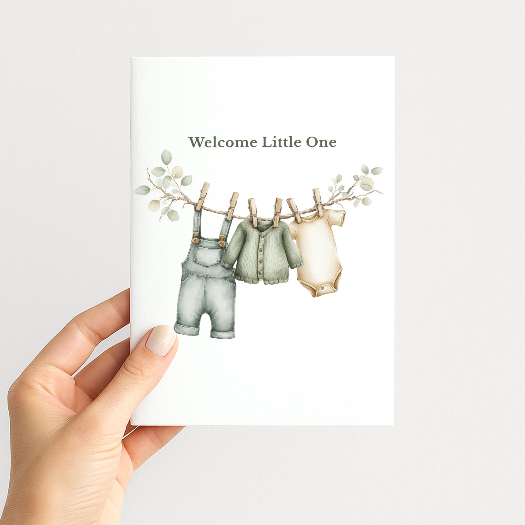 Hand holding a white new baby card illustrated with neutral baby clothes hanging from a eucalyptus-trimmed clothesline.
