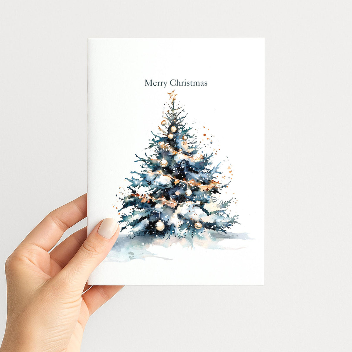 Hand holding a Christmas card featuring a snow-covered evergreen tree with gold ornaments, garland, and a star.