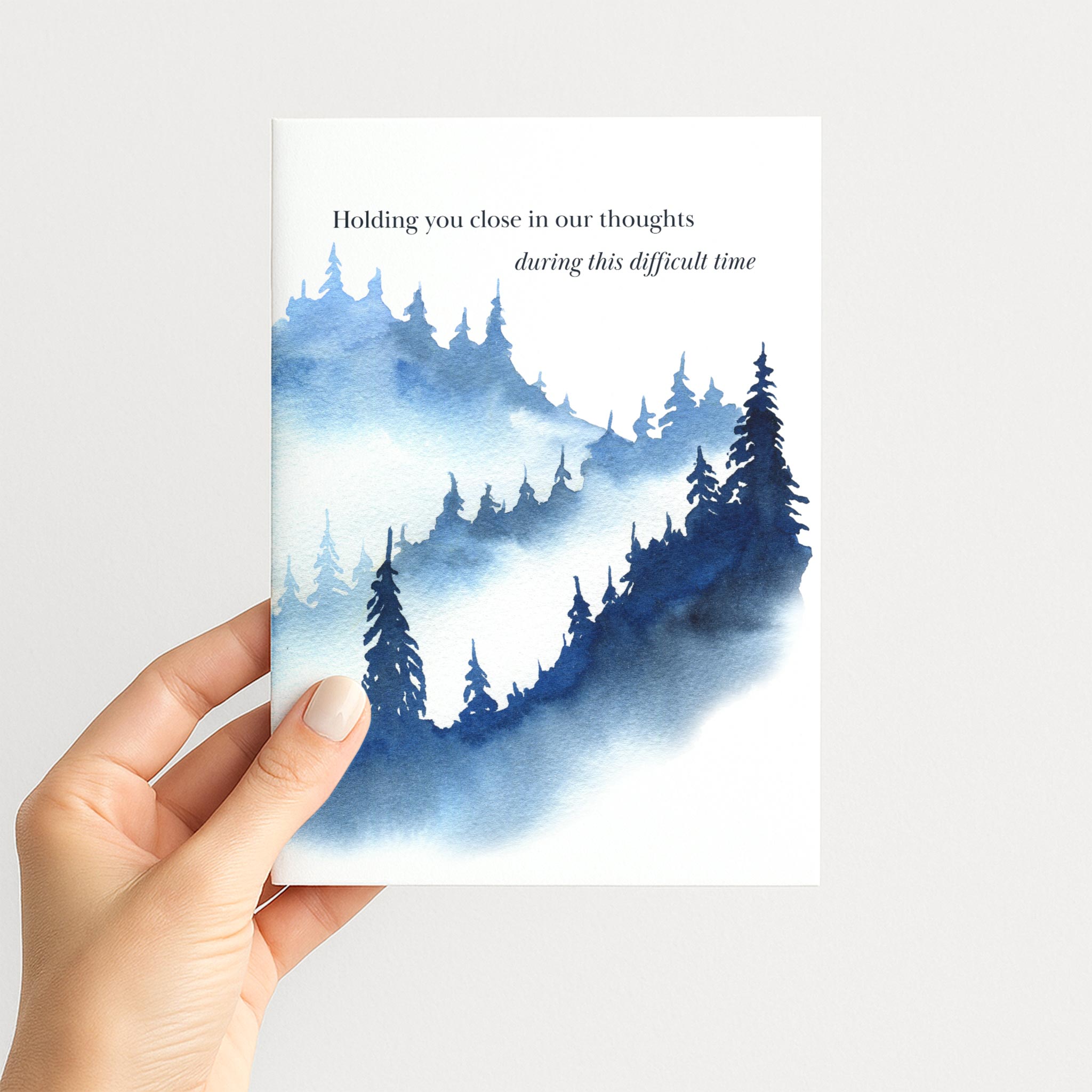 Hand holding a sympathy greeting card featuring misty blue watercolor pine trees and a message of comfort.
