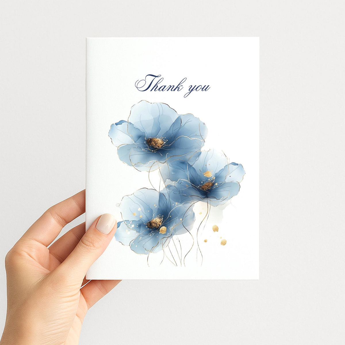 Hand holding a white thank-you card decorated with blue watercolor flowers, gold details, and navy script lettering.