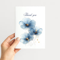 Hand holding a white thank-you card decorated with blue watercolor flowers, gold details, and navy script lettering.