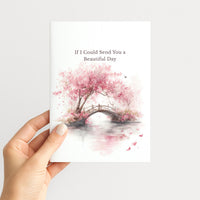 Hand holding a white greeting card illustrated with pink cherry blossoms, a garden bridge, and the message “If I Could Send You a Beautiful Day.”