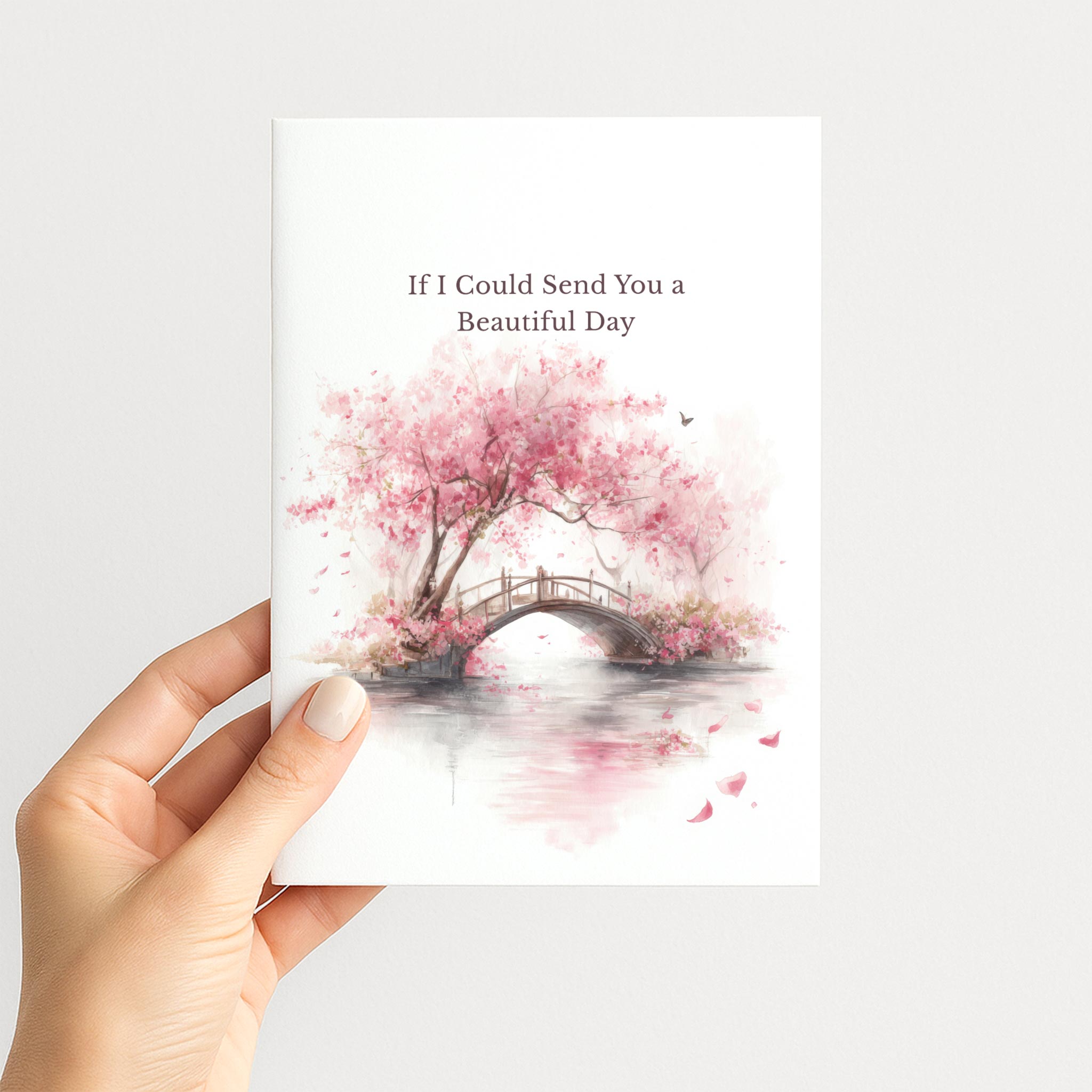Hand holding a white greeting card illustrated with pink cherry blossoms, a garden bridge, and the message “If I Could Send You a Beautiful Day.”