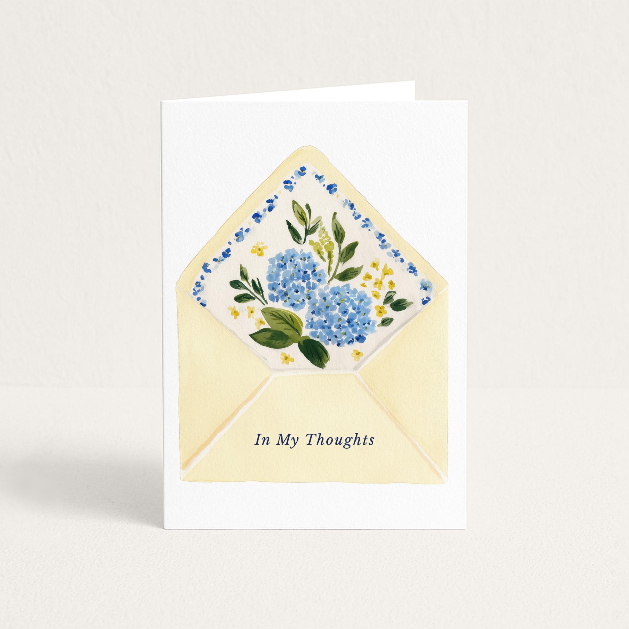 Thinking-of-you greeting card featuring a pale yellow envelope filled with blue hydrangeas and small yellow flowers, with the words “In My Thoughts.”