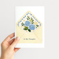 Hand holding a white greeting card illustrated with a pale yellow envelope, blue hydrangeas, green leaves, and the words “In My Thoughts.”