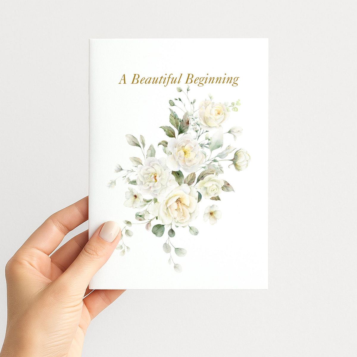 Hand holding A Beautiful Beginning wedding card with delicate white watercolor flowers and gold-toned lettering.