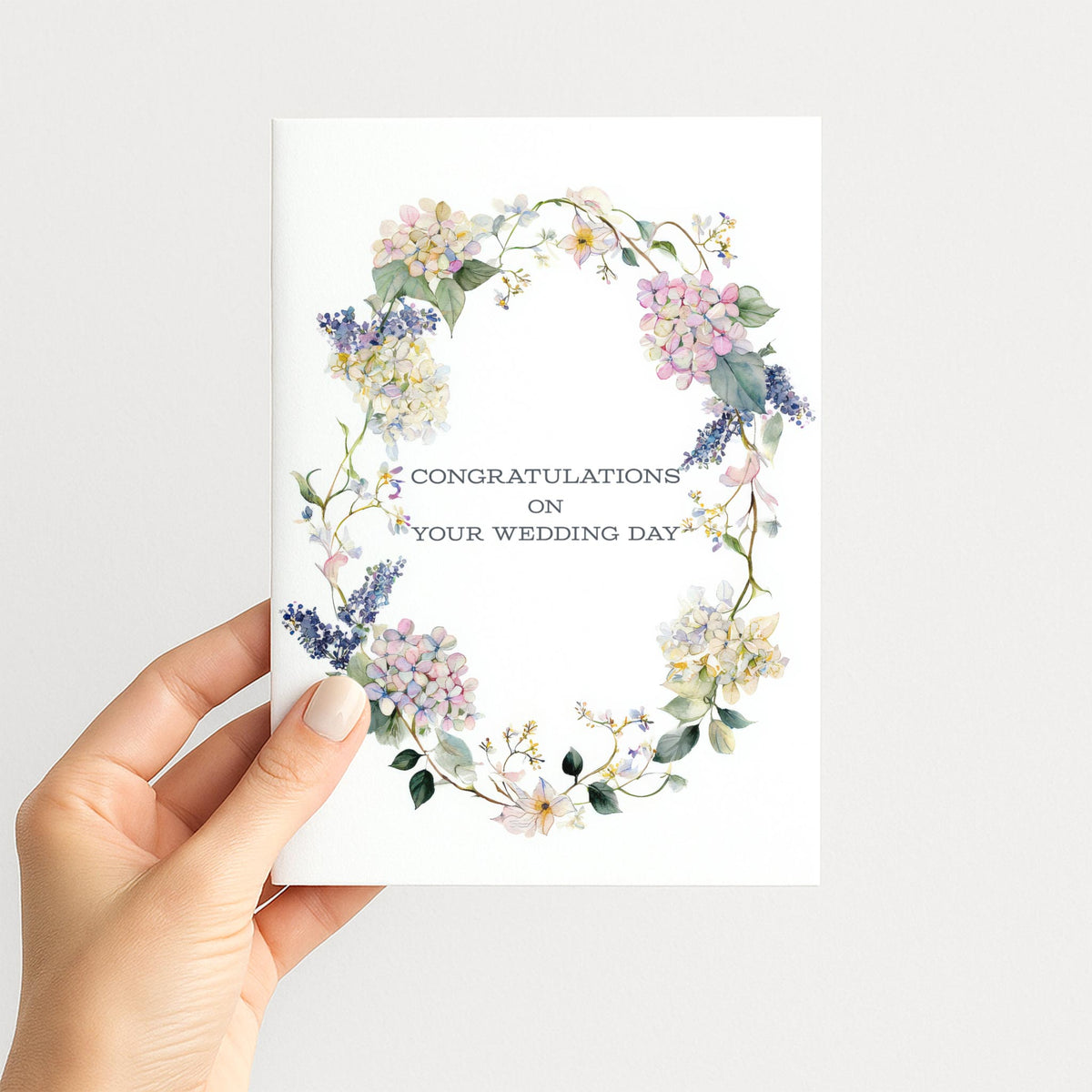 Hand holding a wedding card featuring a pastel watercolor floral wreath with hydrangeas and trailing greenery.