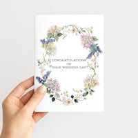 Hand holding a wedding card featuring a pastel watercolor floral wreath with hydrangeas and trailing greenery.
