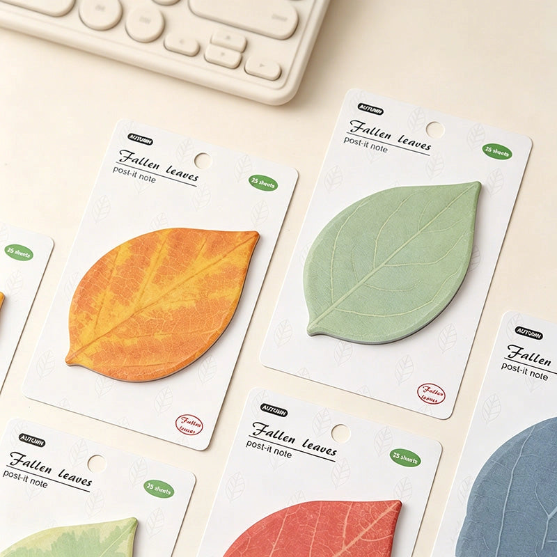 A set of post-it notes shaped like autumn leaves, shown in multiple colors (orange, light green, red, and blue) on their white backing cards, arranged on a light beige desk next to a keyboard.