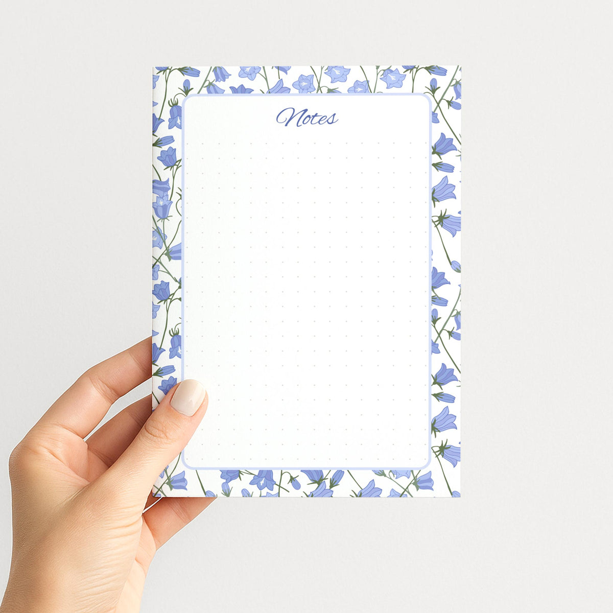 Hand holding the Bluebell Garden memo pad upright, showing its blue floral border, rounded writing panel, and subtle dot pattern.