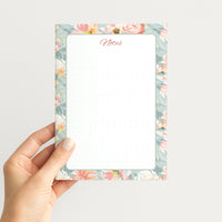 Hand holding the Blush Garden memo pad upright, showing its floral watercolor border, coral Notes heading, and subtly dotted writing area.