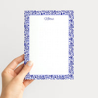 Hand holding the Indigo Botanica memo pad upright, showing its blue-and-white folk floral border, script Notes heading, and dotted writing field.