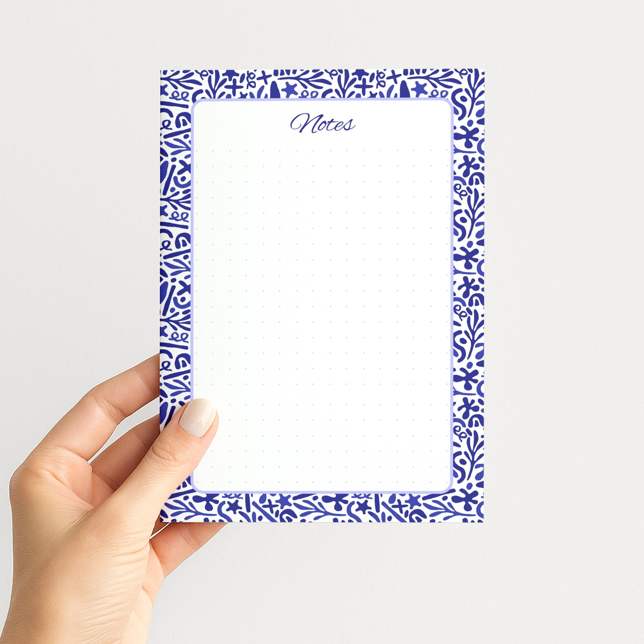 Hand holding the Indigo Botanica memo pad upright, showing its blue-and-white folk floral border, script Notes heading, and dotted writing field.