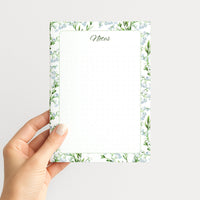 Hand holding the Lily of the Valley memo pad upright, showing its delicate floral border, soft green frame, and subtly dotted writing field.
