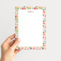 Hand holding the Painter’s Garden memo pad upright, showing its colorful watercolor floral border, script Notes heading, and dotted writing field.